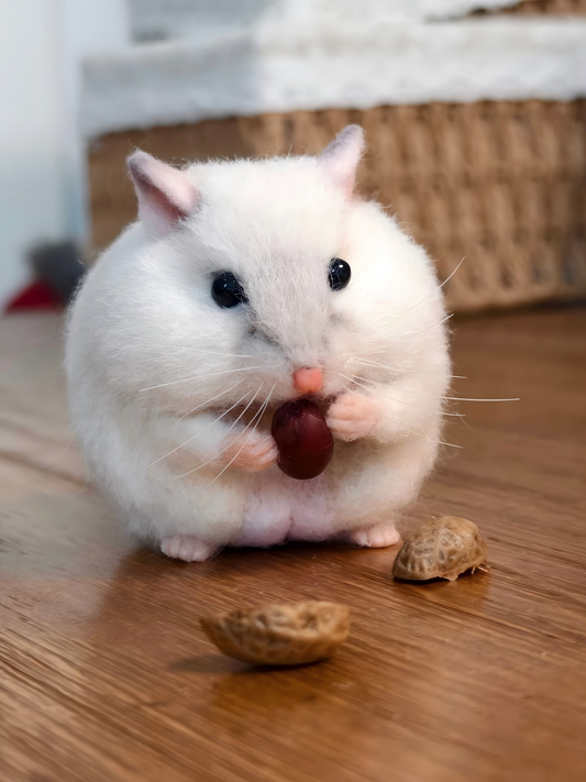 Plush white hamster with dark eyes and pink nose, holding a nut on wood